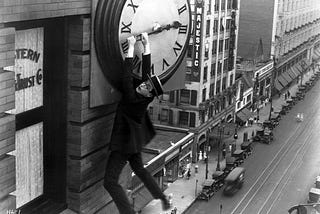 A black and white image of a silent movie. A man with specs and a flat, straw hat hangs off the hand of a clock on the side of a tall building in the city.