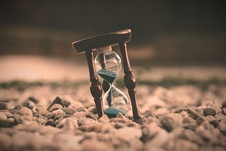 An hourglass with blue sand sits leaning on a bed of rocks, ticking away at this thing we call life.