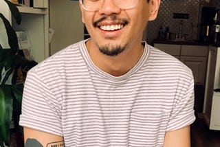 Author Anthony Veasna So, who died last year at age 28, was a rising literary star, smiling broadly, with clear plastic glass frames and a t-shirt with red stripes, with a tattoo of a woman and a text bubble on his right arm (photo by Alex Torres in the LA Times)