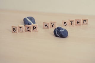 The words “Step by Step” spelled out on Scrabble tiles in front of and behind two blue and white pebbles