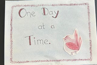 A watercolor bird is pasted to a card that reads “One Day At A Time.”