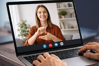 Video call on a laptop screen with a woman in a brown shirt using sign language. Books and plants in the background. Hands typing.