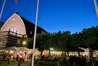A historic, red brick building at twilight with a cozy beer garden next to it brimming with life against a cloudless sky