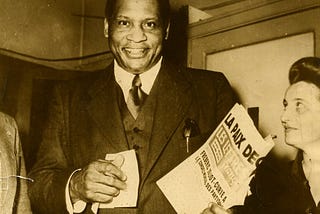 Smiling very tall man wearing a suit and carrying a newspaper. To his right and half cropped from the photograph is a woman smiling and looking upwards towards him