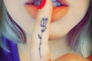 Photo of a finger with a rose tattoo on a woman’s lips as if saying “shhh”