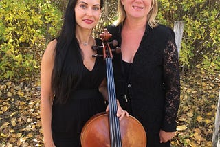 2 women standing outside in formalwear, one holds a cello