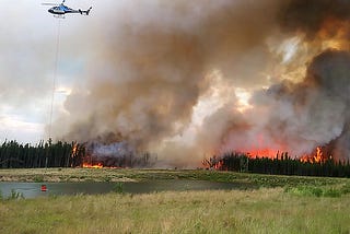 View of the wildfires in Alberta, Canada, showing fire in the timber with massive smoke plume rising in the background, lake and wetland in the foreground, and helicopter filling a drop-bucket with water to drop on the advancing flames. From the Alberta provincial website: https://www.alberta.ca/alberta-wildfire.aspx.
