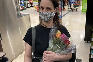 A masked Rebecca holding a bouquet of roses while sitting on her scooter in a grocery store