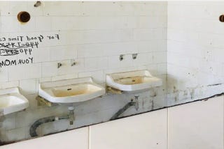 Old, corroded, public bathroom mirror reflecting an image of dirty sinks missing fixtures and the aged, white concrete block wall with lines of graffiti written in black that are reversed in the mirror reflection.