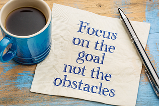 Blue mug, pen, and paper which says “Focus on the goal not the obstacles”