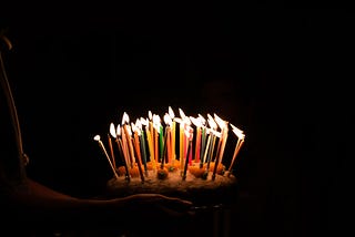 A birthday cake full of lit candles sits is being held while the surrounding area is eclipsed in darkness.