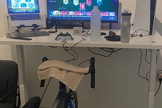 This photo shows a home bike training station setup. The bike is mounted on an indoor trainer stand, and there are two screens on the desk. A game is open on the large screen, while another application or program is running on the smaller one. On the desk, there are a water bottle, towel, and other equipment. A wall-mounted shelf holds decorative items, small plants, and framed photos. On the right side, there’s a large white device, likely a gaming console.