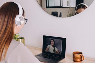 A feminine-presenting white person with long light brown hair sits at a shelf desk mounted to a white wall. She has white headphones on and leans toward a laptop that’s on the desk. Another feminine-presenting person with dark hair is on the laptop screen, indicating a video conference with a career discovery coach or counselor.