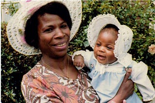 Black woman in a hat holding a small Black baby wearing a bonnet.