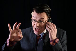 Middle-aged white man with slicked back short black hair against a black background, mouth agape, appearing to argue on the phone while showing the A-OK or “asshole” symbol with his right hand. He’s wearing a black suit jacket with a red plaid shirt and blue tie, with wire-rimmed glasses.
