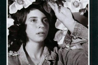 This is the album cover for Cat Power’s Moon Pix LP. It is a black and white picture and features a woman in a denim jacket surrounded by flowers. The text is in red.