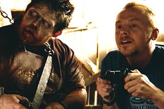 Simon Pegg as Shaun and Nick Frost as Ed in Shaun of the Dead are playing video games on a sunny day as rays of light lighten the area behind them. Shaun is smiling. Ed is a zombie.