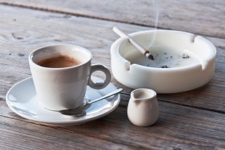 A cup of coffee and an ashtray with a smoldering cigarette on a wooden table