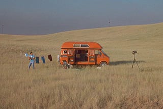 A man stands in a field next to the van he’s living in, holding a football, and recording himself with an old camcorder on a tripod.