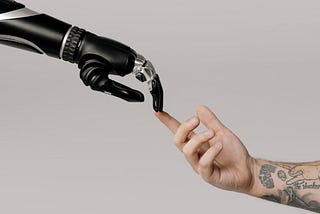 Two hands reaching for each other: the one on the left is robotic, the one on the right tattooed and human