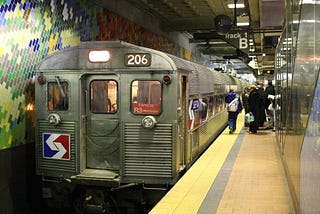 Subway train approaching with yellow caution paint at the end of the platforms. A few people are waiting for the train.