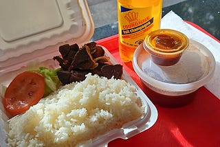 A Haitian meal of rice, deep-fried beef, a soda in a bottle, and a bowl of beans and sauce with some pan drippings