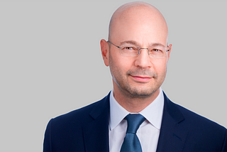 jason colodne wearing a blue jacket and a blue tie with glasses in front of a gray background