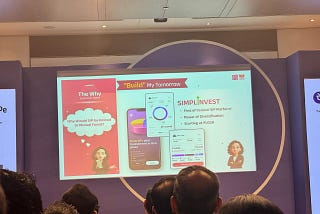 A presentation slide for Simplinvest displayed on a large screen at Global Fintech Fest 2025, with an audience visible in the foreground. The slide is titled “Build My Tomorrow” and features the Simplinvest logo alongside three key highlights: “First of its kind SIP Platform,” “Power of Diversification,” and “Starting at Rs 1,000.” The left panel poses the question “Why should SIP be limited to Mutual Funds?” under the heading “The Why: Customer Need.” The center shows mobile app screens illustr