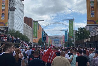 A large crowd of people can be seen walking down Olympic Way towards Wembley Stadium, whose iconic arch can be seen in the distance. The sky is filled with a number of large clouds. England flags can be seen draped over railings along the walkway and large Euro 2020 branded banners flank the walkway on either side.