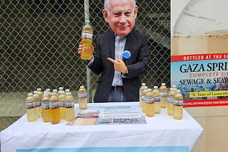 A man wearing a suit and a Netanyu mask points to a plastic bottle filled with yellow-colored liquid and labeled “Gaza Springs” as he stands behind a table with more bottles and some literature, next to a large poster showing an enlarged bottle with the label saying “Bottled at the source — GAZA SPRINGS — Complete Siege Sewage and Seawater — ‘A Taste of Genocide.’”