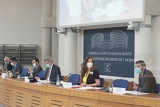 A photo of the bench of the European Court of Human Rights. Sat at it are 6 individuals, all wearing face masks