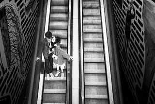 A black and white photo shows a twin set of escalators, with a hetero-presenting couple on the left, about to kiss.