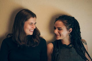 Two women in their twenties sit against a beige wall. One is a white woman with long dark hair, and the other is a light-skinned black woman with dreadlocks. They are both smiling.