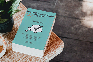 A turquoise book cover with the title “The Evoluationary OKRs Playbook” with the Thinkydoers logo, a black outlined, white thought bubble, with a large black checkmark in the middle.