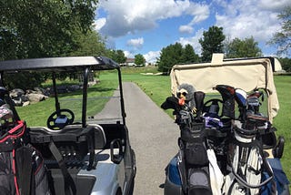 Photo of the back of two golf carts with 3 sets of golf clubs. The Carts are at the tee blocks of the par three number nine hole facing the green.