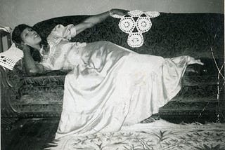 A picture of an African American man in a white wedding dress. The dress has a soft white sheen. Sweets is lying on the couch in a reclined pose. Sweets’ right arm props her head up, while her left arm is extended touching a crocheted doily on the back of the couch.