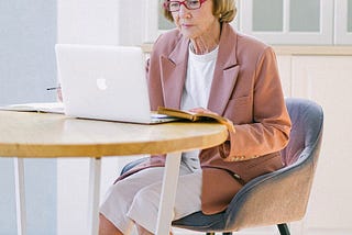 An older woman sitting at the kitchen table and looking at her laptop.