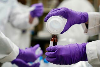 Lab worker in purple gloves pouring a liquid from a container into a smaller vial