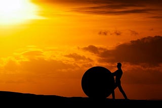 A sunset is behind a man’s silhouette. He’s pushing a large boulder up a hill.