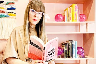 A woman reads the dark satiric wellness influencer novel Self Care by Leigh Stein in a pink room.