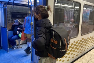 A picture of our team member, Mia, boarding the subway in Pittsburgh. She has dark hair in a bun, and is wearing a black puffy jacket, a black and brown backpack, and green pants.