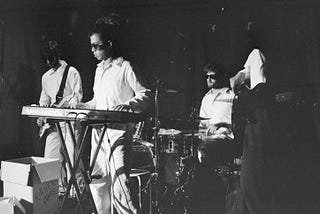A black and white photo of performers playing musical instrument on stage at the Edinburgh fringe. They are all wearing black sunglasses and white clothes.