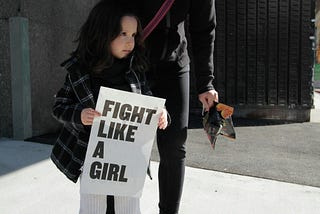 girl holding fight like a girl-printed paper beside woman holding plastic pack