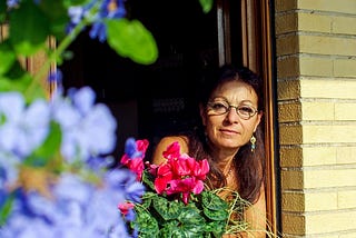 Middle-aged brown-skinned woman in glasses and long brown hair peeks out of an open window or door at vibrant pink and periwinkle flowers, against a yellow brick building. She’s wearing dangling earrings and appears to have pigtails.
