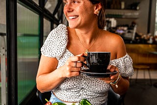 woman looking out the window smiling holding a cup of coffee or tea with a salad on the table in front of her