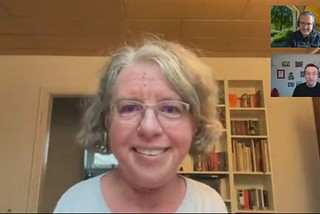 A screenshot from a podcast recording featuring Katrin Langensiepen as the main speaker. She is a middle-aged woman with short, curly gray hair, wearing glasses and a light-colored top, smiling at the camera. Behind her is a bookshelf filled with books and decorative items. In the top-right corner, two smaller video windows show the podcast hosts; one is wearing glasses and a dark jacket, with a blurred outdoor background, while the other is in a room with framed pictures on the wall, wearing a black shirt.