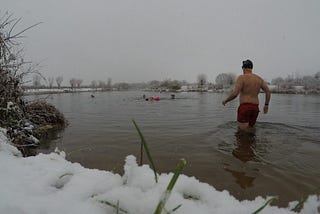 A man wading into a river, away from the camera, snow on the ground in the foreground