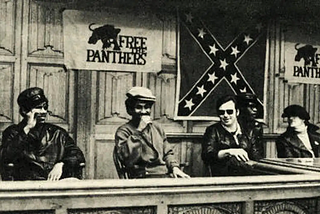 Two Black Panthers sit with two Young Patriots at a table. Free the Panther banners and a confederate flag hang behind them.