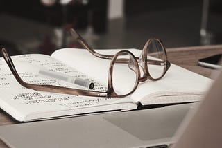 Glasses and a pen on a journal in front of a laptop