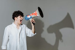 A young man with curly hair yelling through a megaphone with his shadow behind him doing the same.